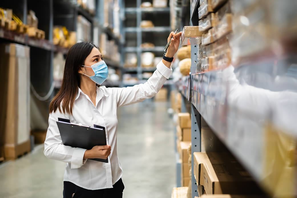 A woman wearing a blue surgical mask and white shirt carries a clip board and takes inventory in a stockroom