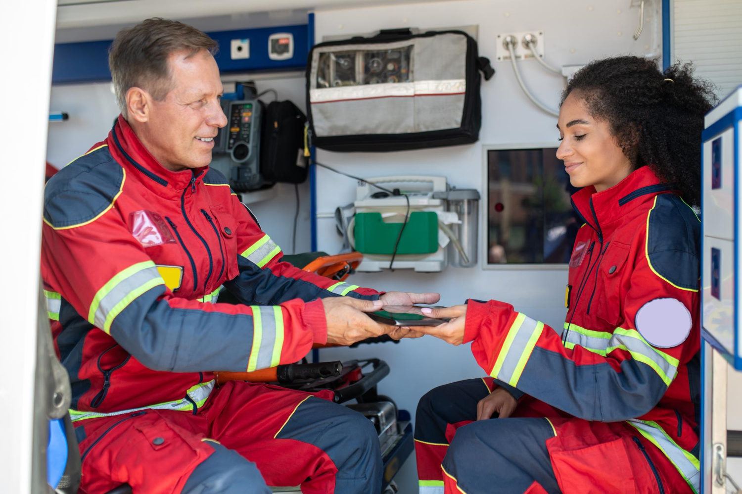 Two EMS workers sitting in an ambulance, wearing red and gray EMS uniforms. A male paramedic hands a tablet to a younger female paramedic.