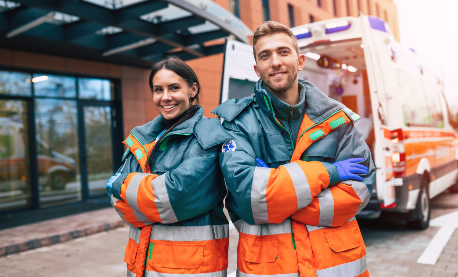 Two paramedics stand in front of an open ambulance bay outside of the hospital. The male and female EMS professionals pose at the camera with their arms crossed, smiling, wearing orange and blue high-visibility jackets.