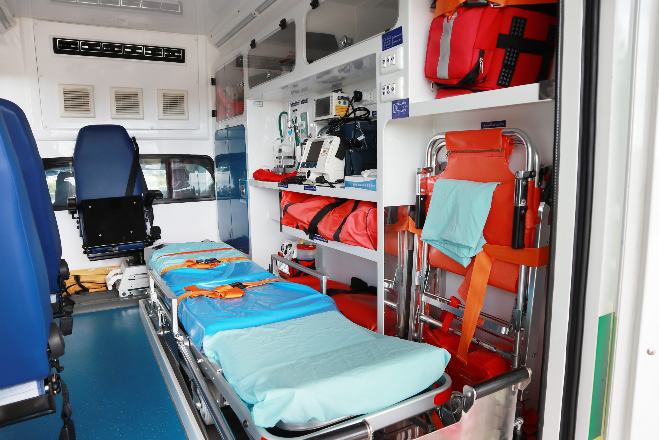 The interior of an ambulance, showing a medical stretcher, seats, and a wall packed with medical and rescue supplies.