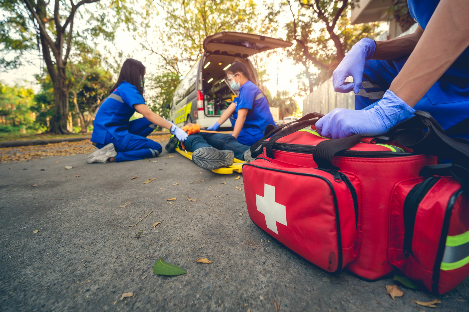 A group of paramedics treat a patient laying on a backboard on the ground while another paramedic holds a first aid supply bag.