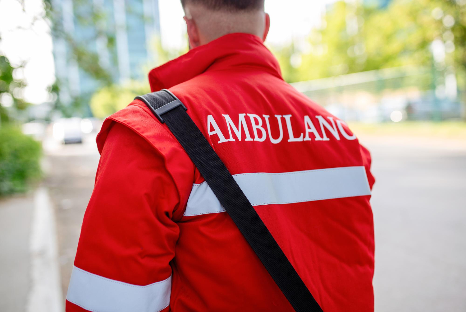 A paramedic stands with his back to the camera. He's wearing a red jacket with the word "ambulance" on the back, along with high-vis strips, and a medical bag strap over his shoulder.