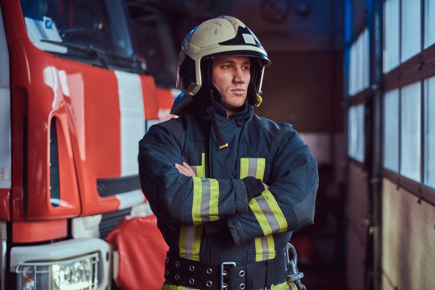 A male firefighter stands inside a firehouse, in front of a red fire truck. He's wearing a protective jacket and helmet, standing with his arms crossed, looking away from the camera.