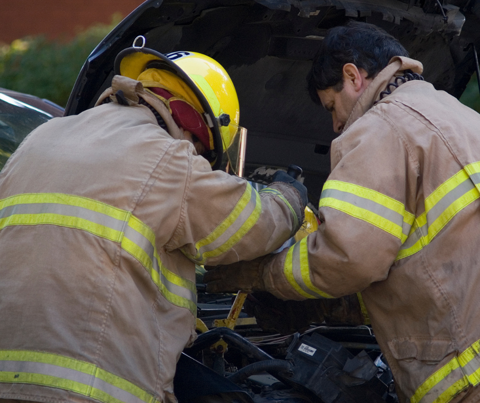 Two firefighters in turn-out gear work under a car’s hood.