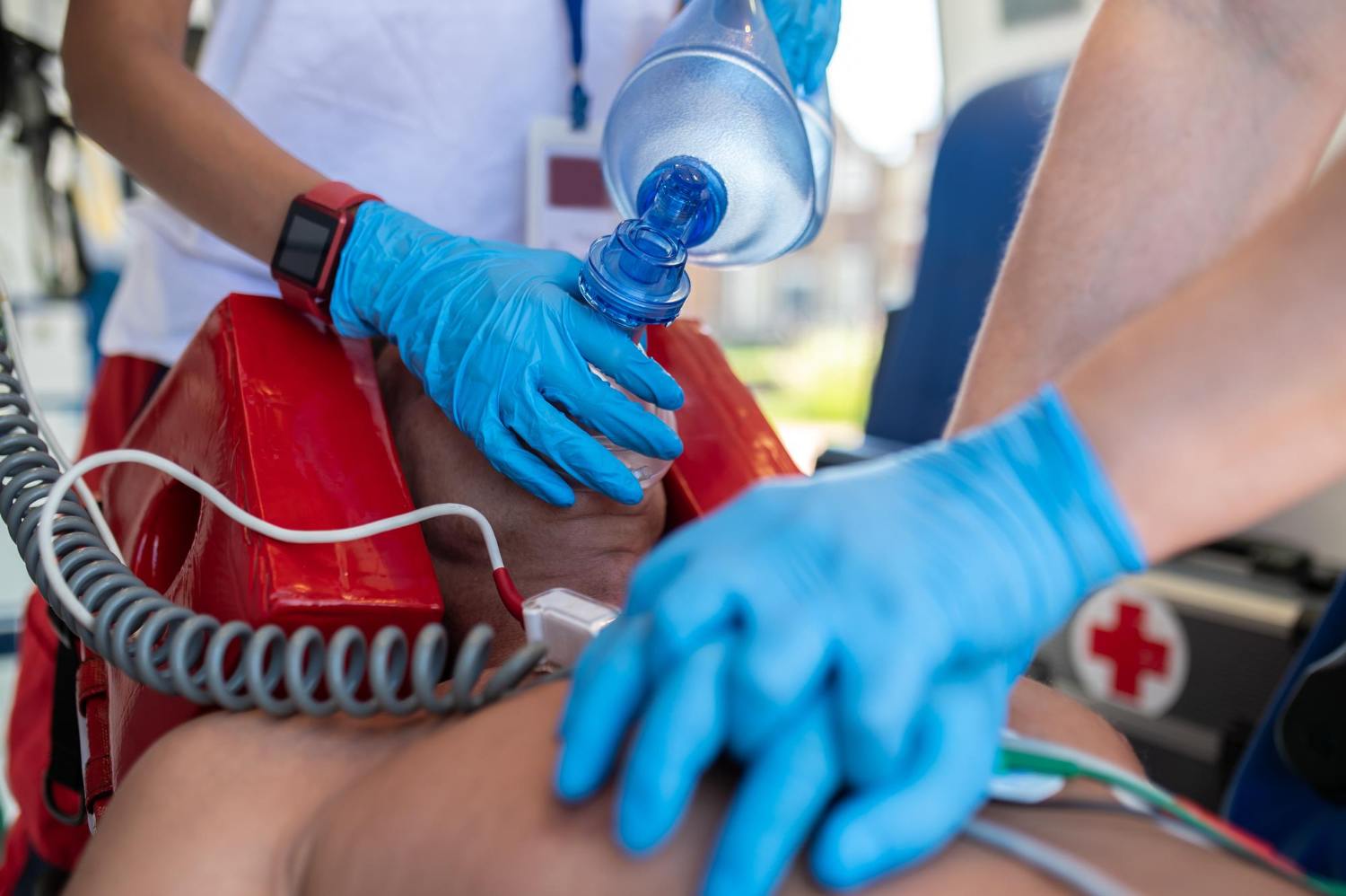 An EMT performs CPR on a man laying on a stretcher in an ambulance, while another EMT holds and oxygen mask over the patient's face.