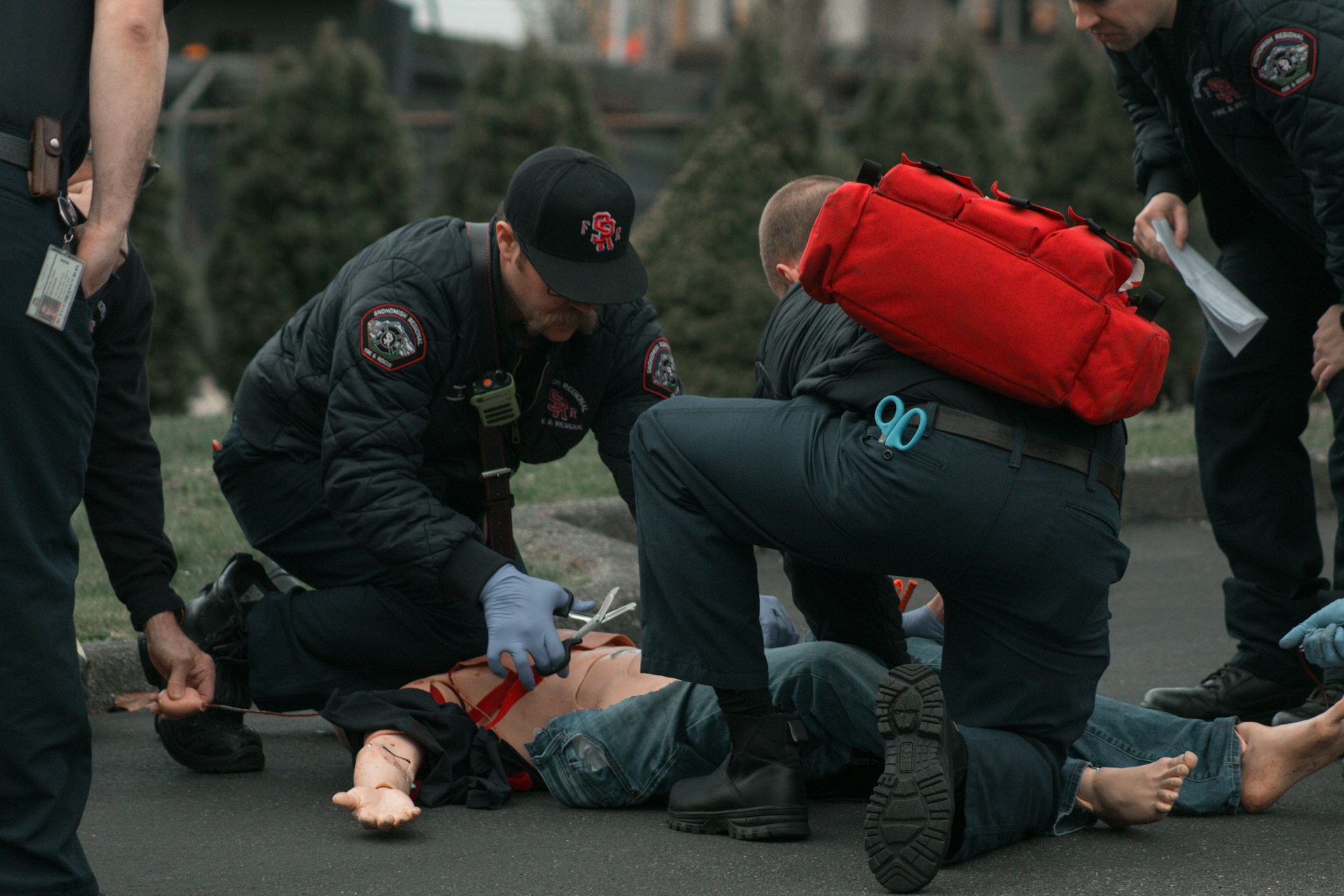 A pair of first responders in EMS uniforms work on a paramedic dummy laying on the ground.